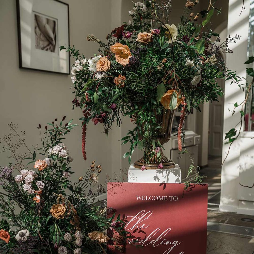 Large maroon wedding welcome sign with white calligraphy surrounded by dried wildflower wedding flower arrangments in large gold vases 