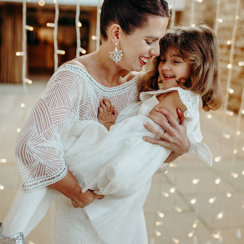 Bride holds flower girl with fairy lights curtain twinkling behind 