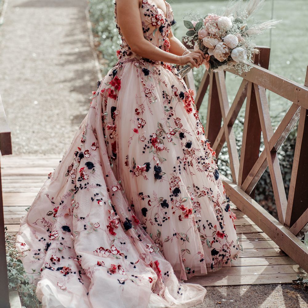 Bride stands on wooden bridge whilst wearing floral appliqué wedding down carrying floral bouquet