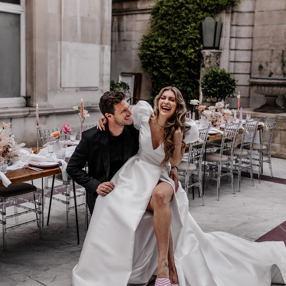 Bride in a front split Love In Lace Bridal gown and pink mules sitting on her husbands lap at their luxury wedding 