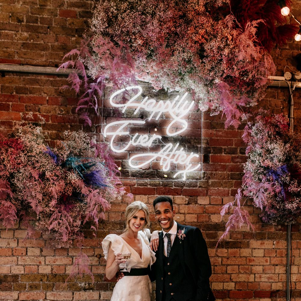 Portrait of the bride in a ruffle dress and groom in a navy wool suit at The Loft Studios under a neon Happily Ever After sign surrounded by a pink gypsophila flower cloud 