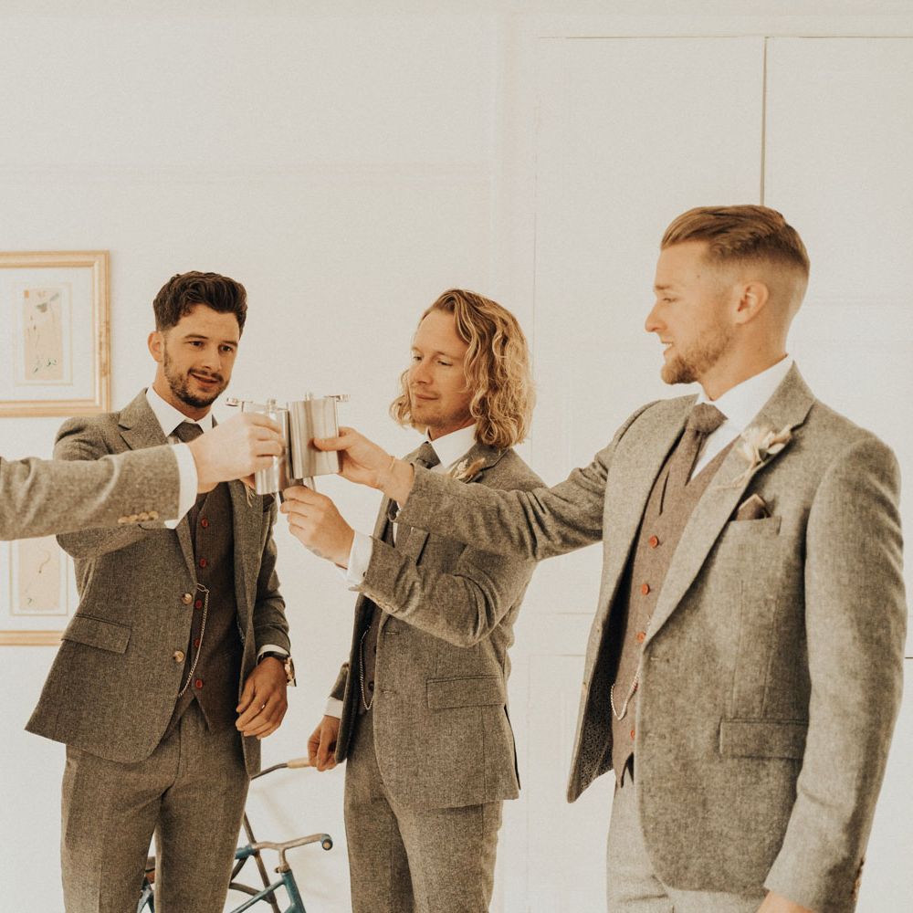 Groom and groomsmen all in sage green three piece suits raise their silver hip flasks to one another before wedding at Anran Devon