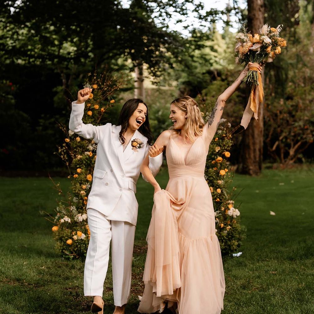 Bride in a white suit and blush pink wedding dress exiting their outdoor wedding ceremony as wife and wife 