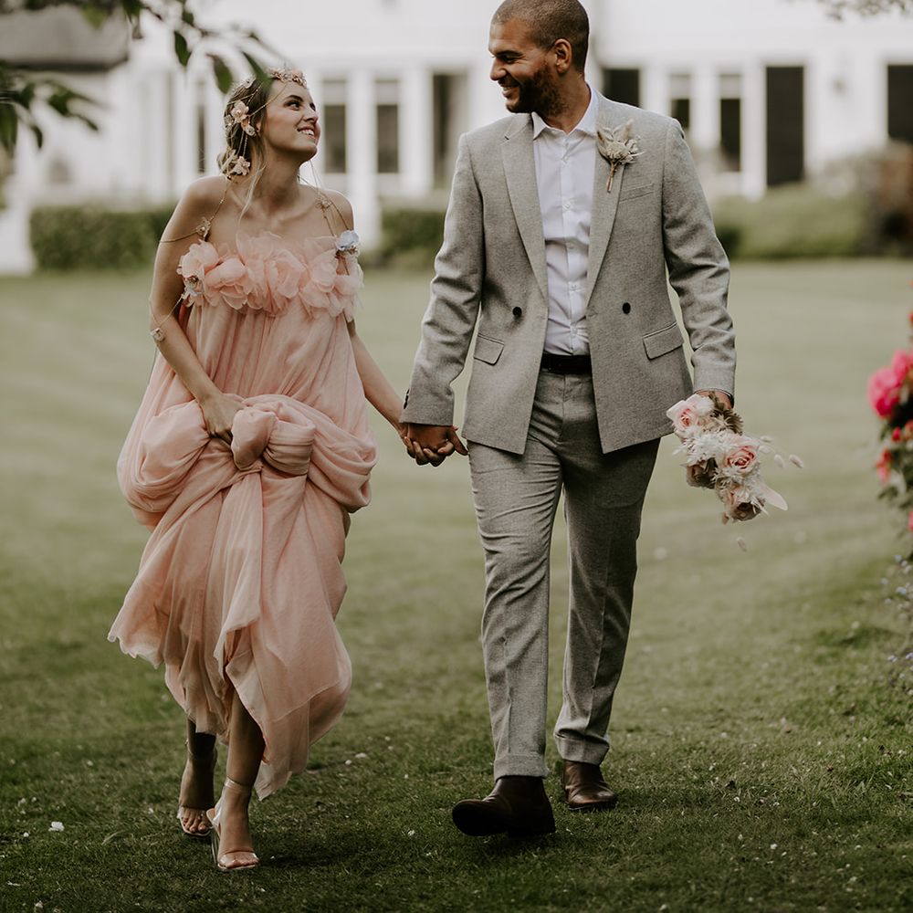 Groom in a light coloured suit holding hands with his bride in a blush pink jumpsuit in front of Waresley Park Estate French Chateau house 