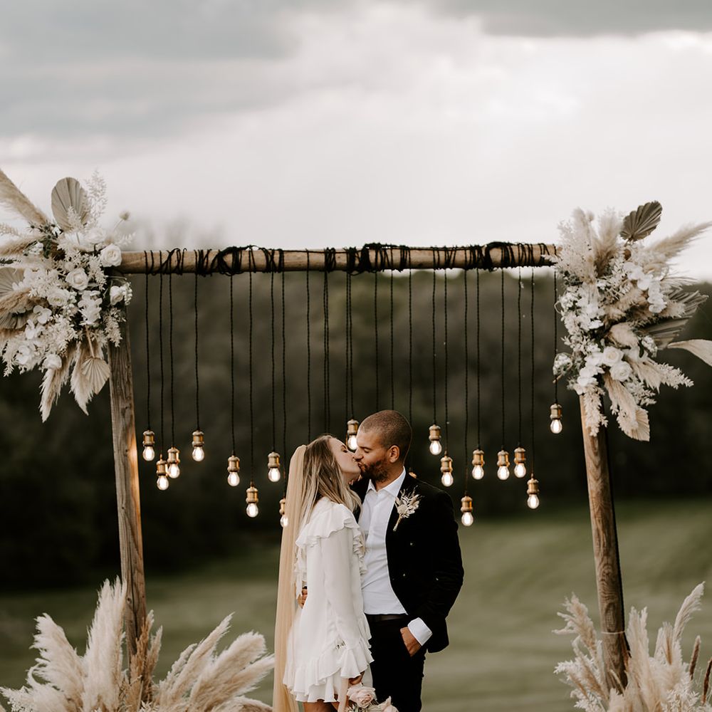 Bride in a short wedding dress clear shoes and long veil kissing her husband at the wooden frame altar decorated in pampas grass flower arrangements and hanging Edison bulbs