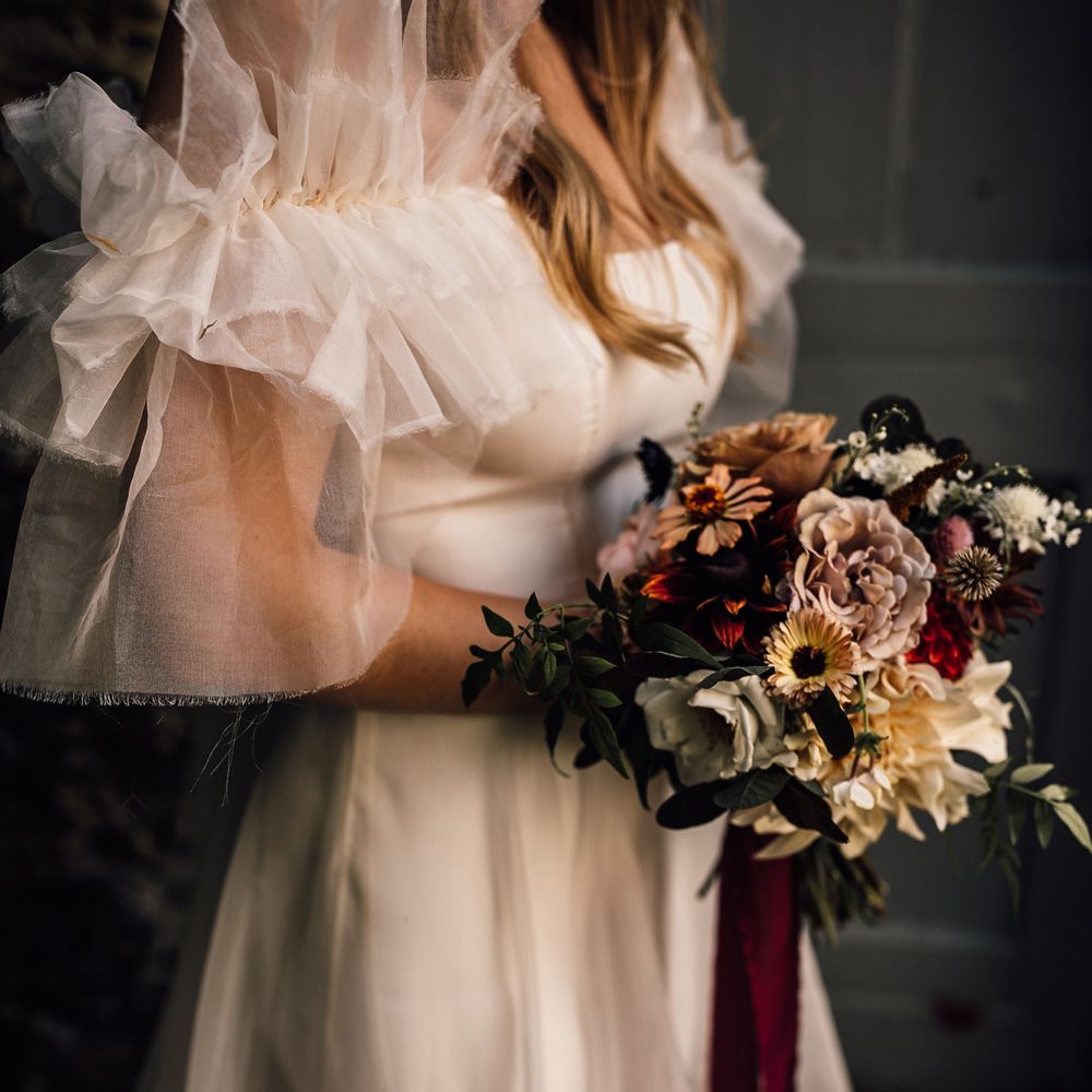 Bride in white Halfpenny London Mayfair dress with ruffle sleeves holding mixed bridal bouquet with burgundy ribbon at Cornwall wedding