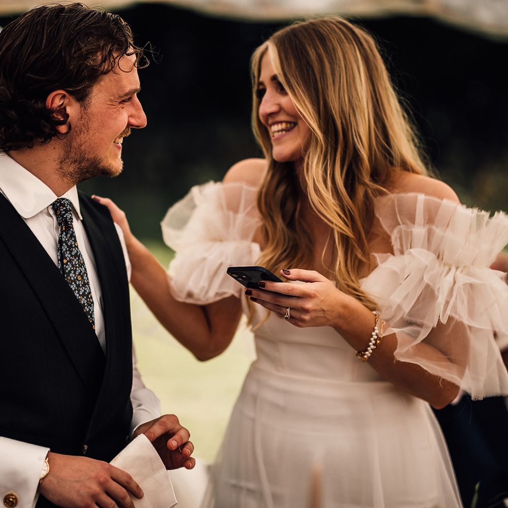 Groom in white shirt, blue waistcoat and blue patterned ties smiles at bride in white Halfpenny Mayfair London dress as she makes speech during garden marquee wedding reception in Cornwall 