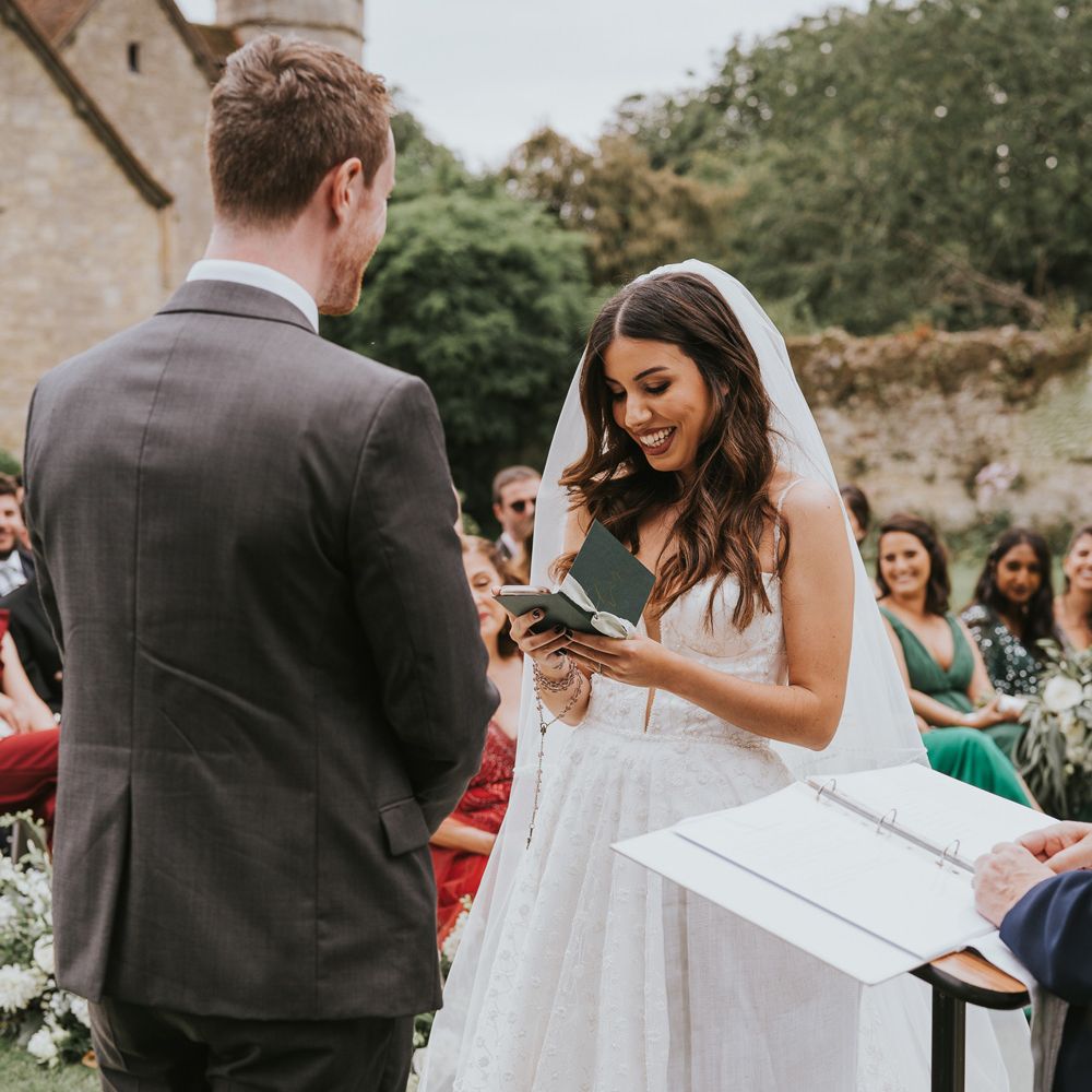 Bride in Pronovias wedding dress and veil reads vows during outdoor wedding ceremony at Notley Abbey as she stands opposite groom in grey suit 