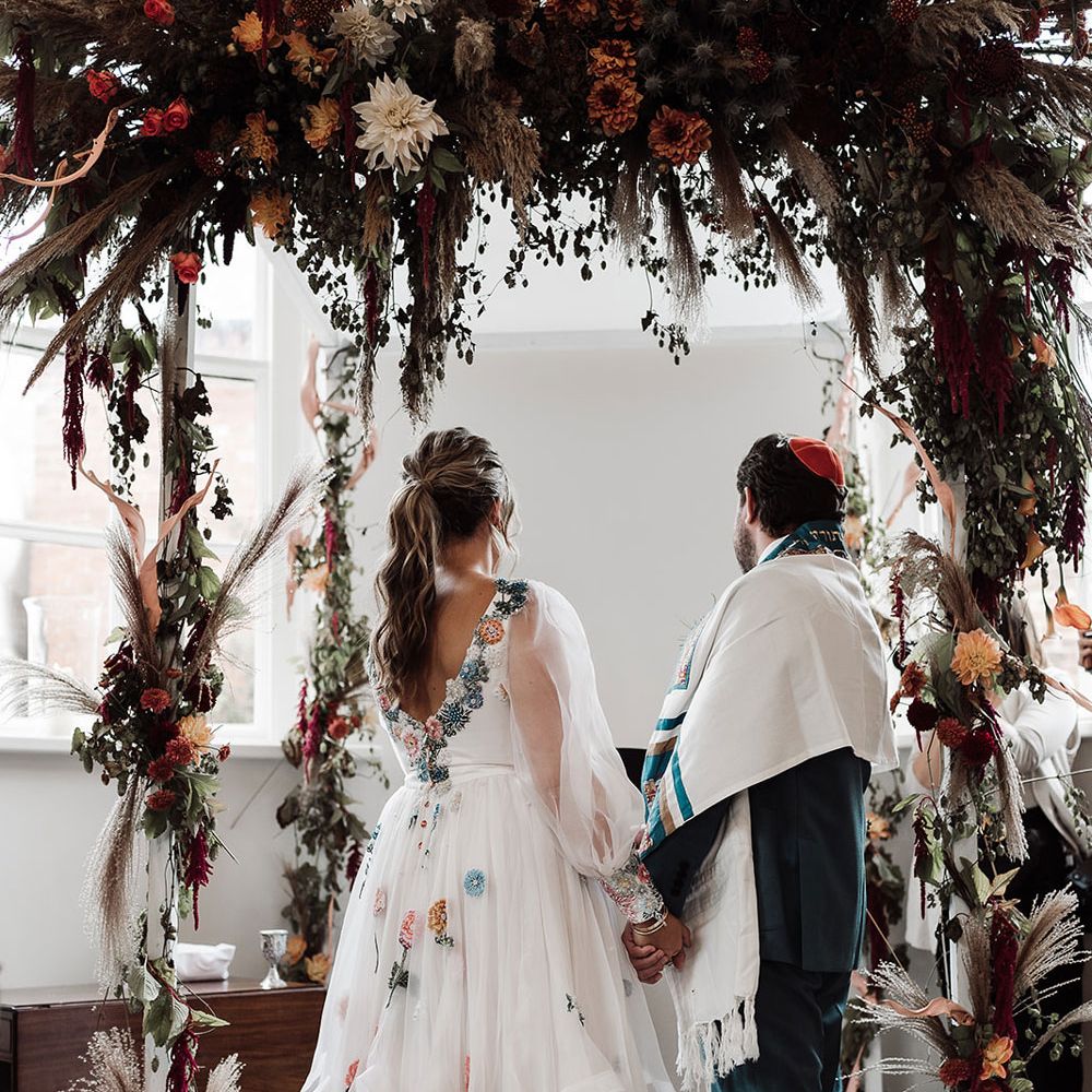 Bride in low back embroidered Victoria Sanders wedding dress with train stands with groom in green suit and red kippah under wedding arch with dried flower cloud 
