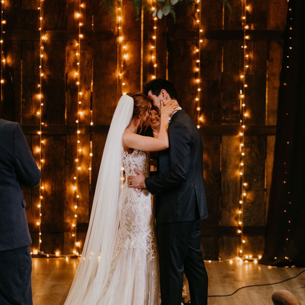 Bride in chapel length veil and lace wedding dress hugs groom in dark suit during fairy lit wedding ceremony in barn