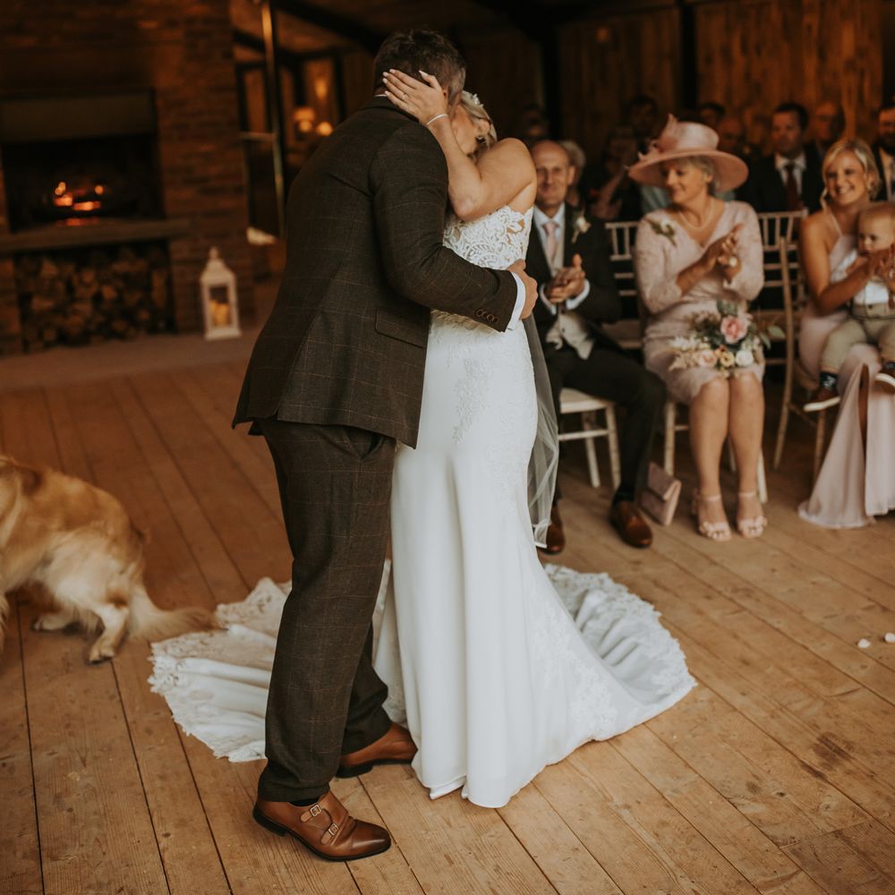 Bride in lace wedding dress with train kisses groom in dark brown suit during rustic wedding ceremony at the Hidden River Barn wedding venue