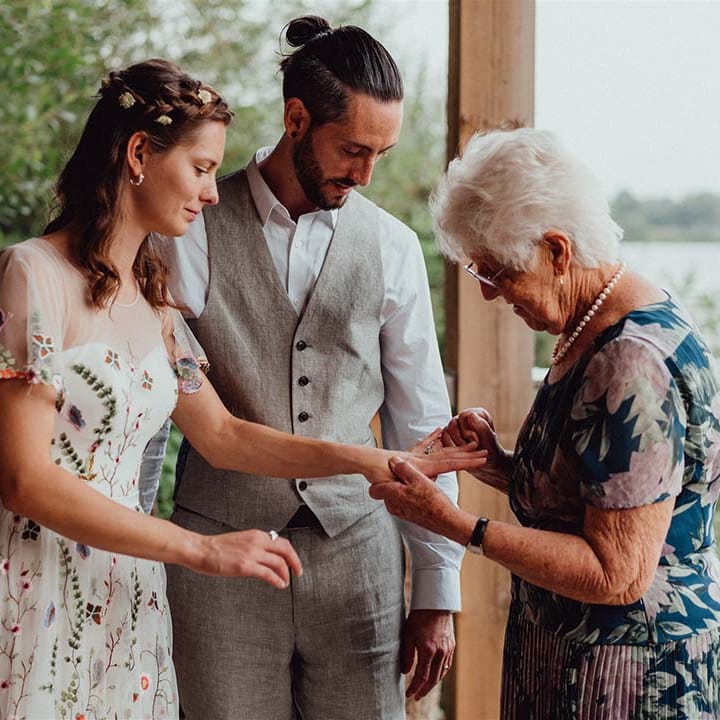 Wedding guest takes a closer look at the bride's wedding rings in floral embroidery wedding dress with groom in white shirt and grey waistcoat 