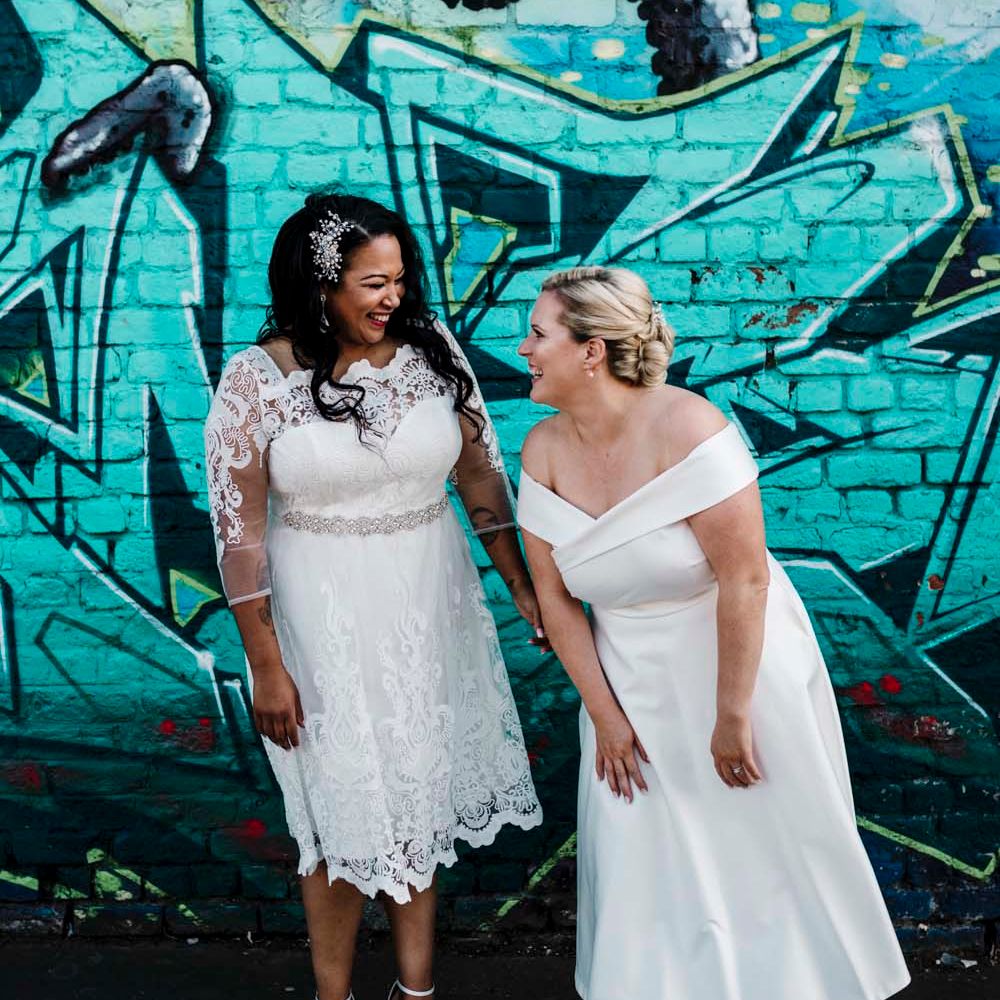 Lesbian wedding with two brides in tea length wedding dresses laughing in front of graffiti art in Liverpool 