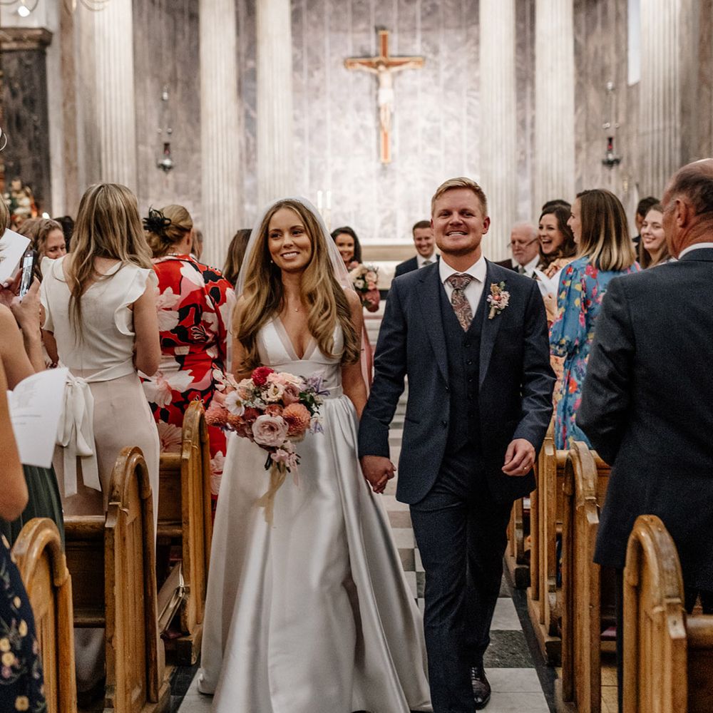 Groom in a three piece navy blue wool suit holding hands with his bride in a Karen Willis Holmes wedding dress as they descend up the aisle as husband and wife 