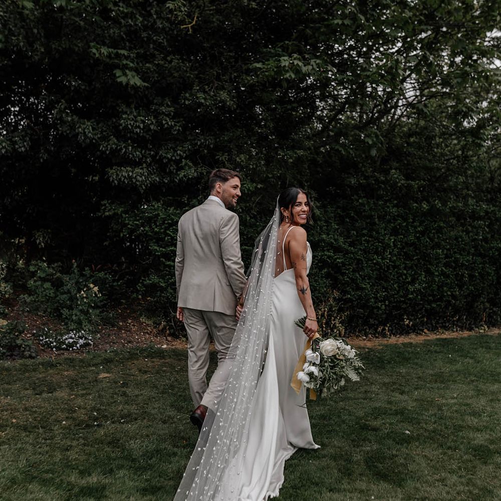 Groom in a beige wedding suit holding hands with his bride in a fitted wedding dress and long pearl veil 
