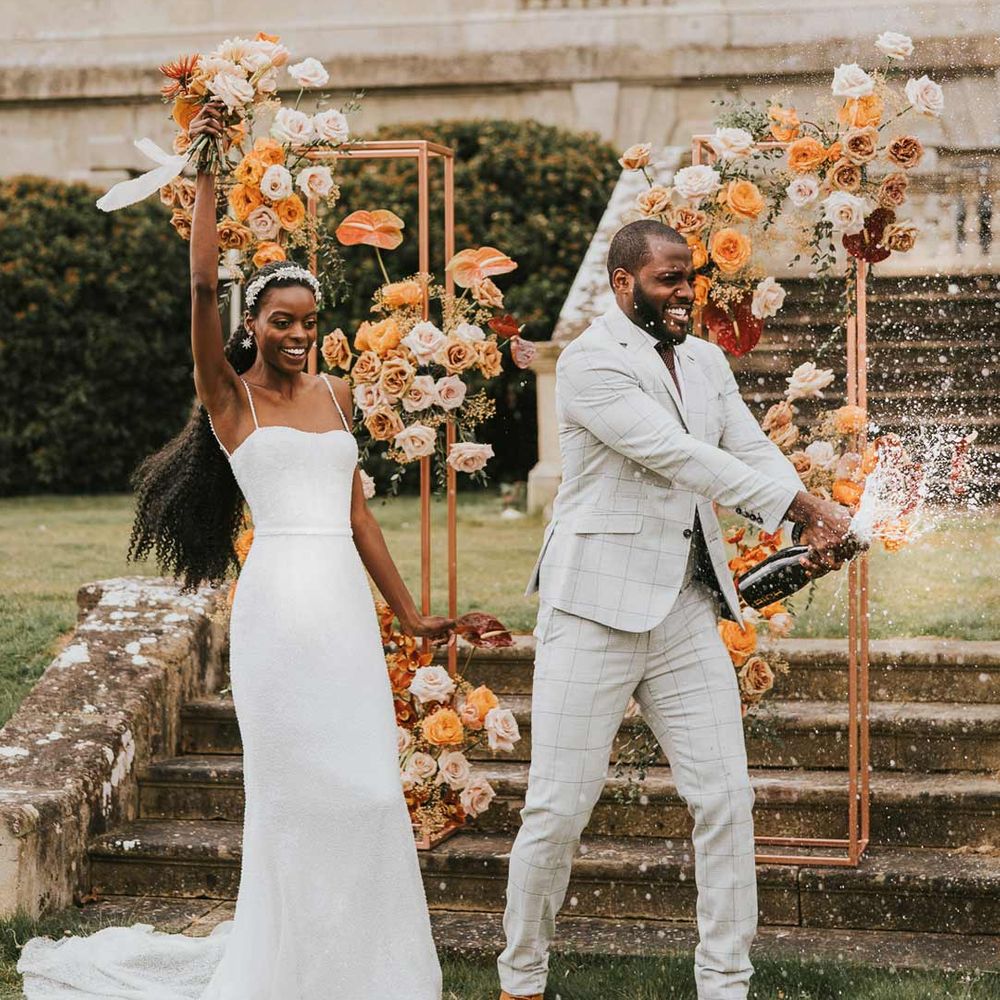 Bride in pearl strappy wedding dress and groom in grey checkered suit doing champagne pop