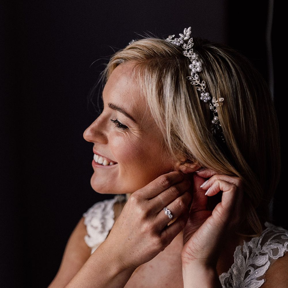 Blonde bride puts in her earrings wearing a gold leaf and pearl headband with a three stone engagement ring 