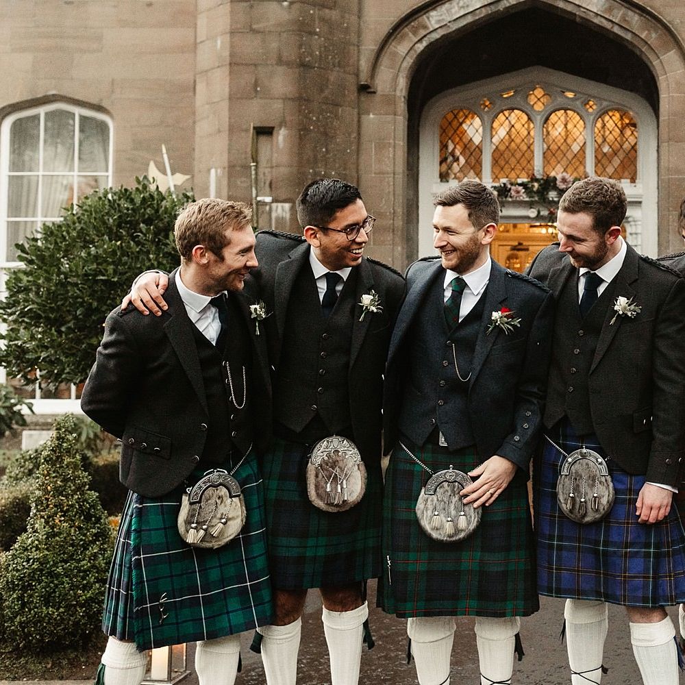 Groom stands with his groomsmen who wear kilts and three piece suits and floral buttonholes 