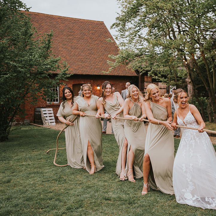 Bride in floral lace wedding dress and bridesmaids in sage green dresses play tug of war 