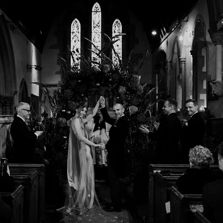 Bride in satin red wedding dress with cape and groom in three piece suit with red tie holding hands at winter wedding alter