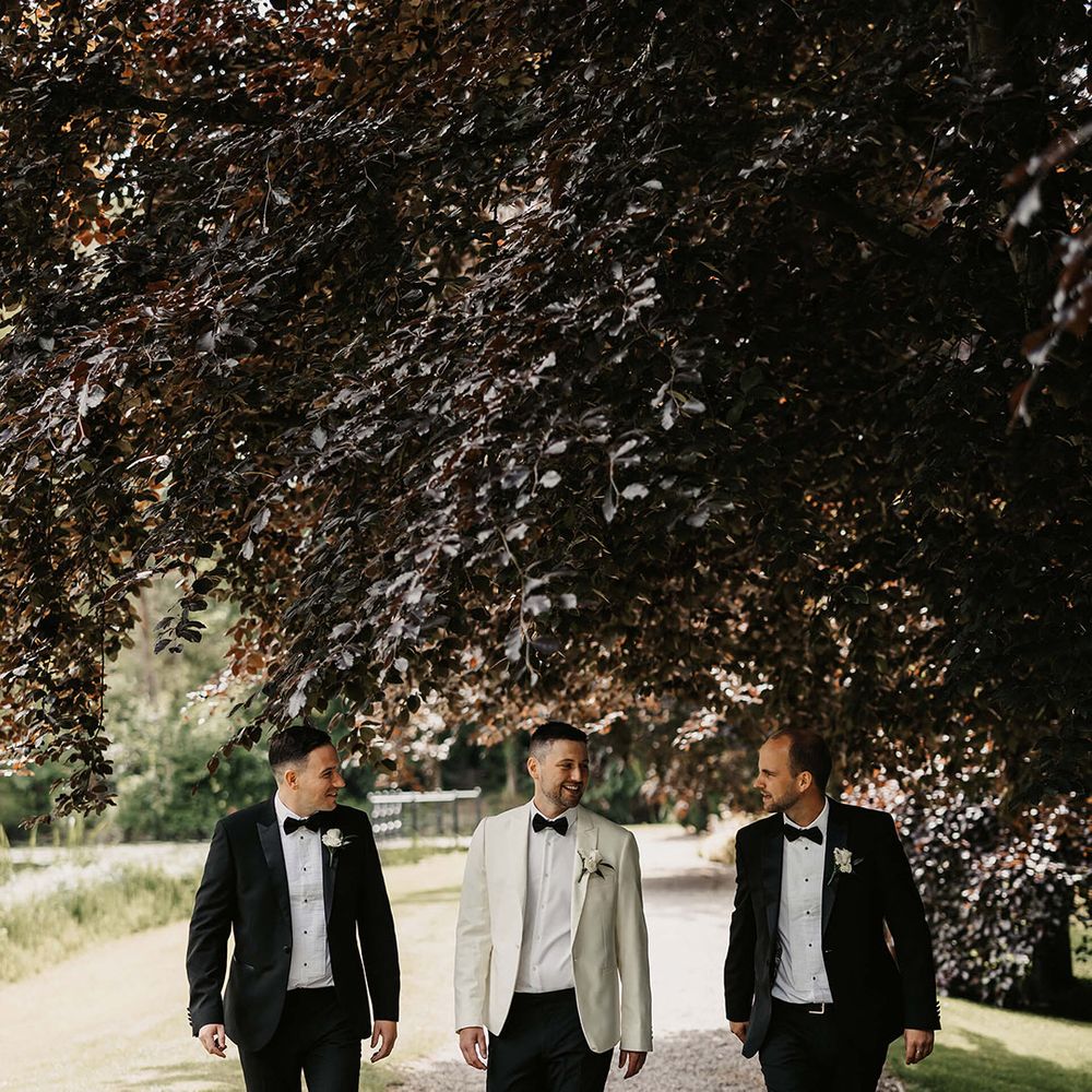 The groom in a white tuxedo walks with two of the groomsmen in black tie all wearing white rose buttonholes 