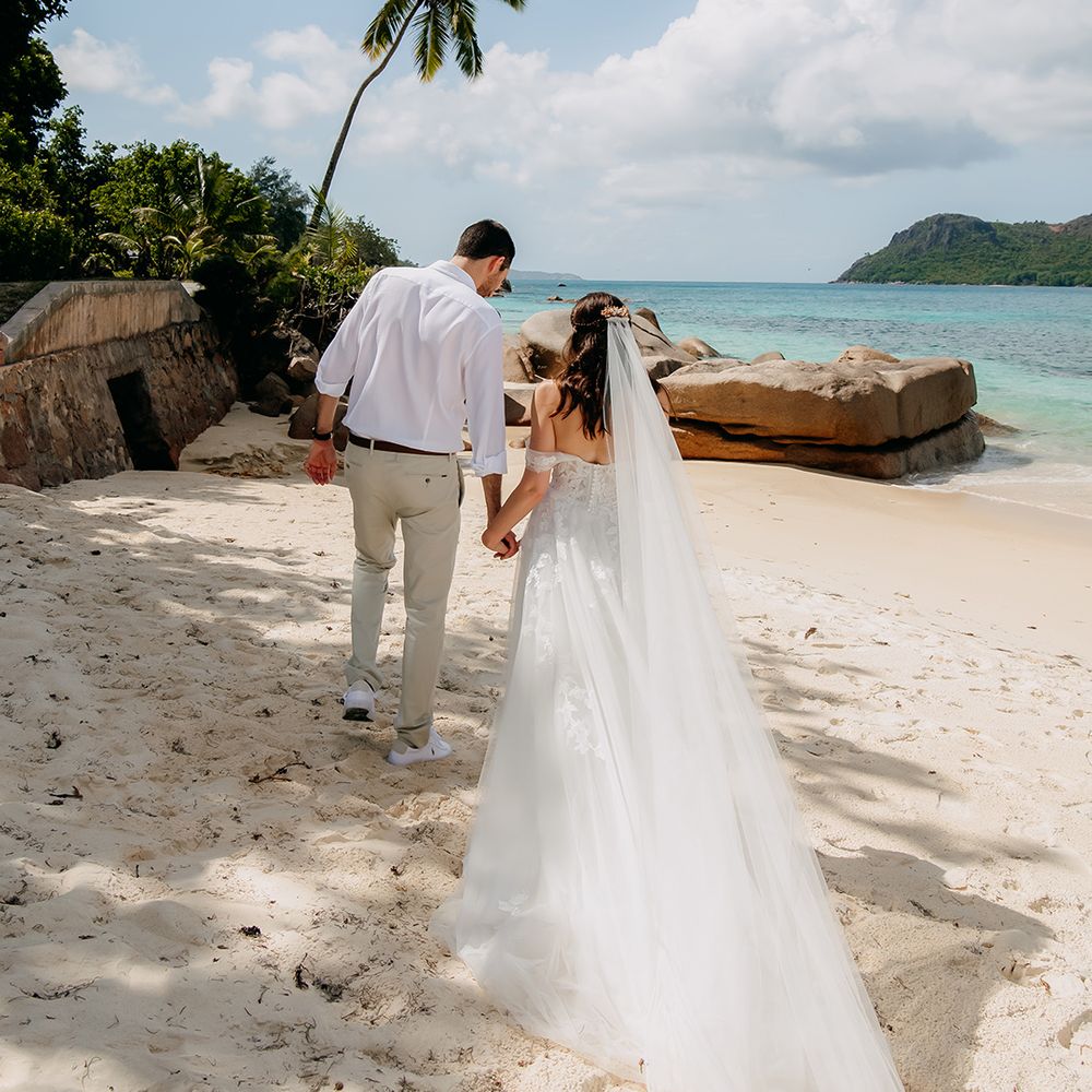 Bride in off the shoulder lace wedding dress with sweetheart neckline and lace floral details and cathedral length veil holding hands with groom in crisp white shirt, light coloured chinos, tan leather belt and white trainers walking on the beach in Seychelles