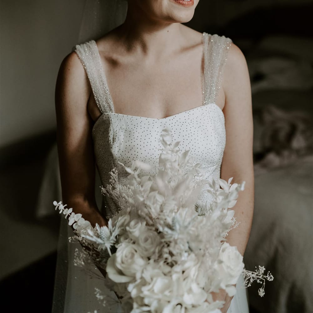 Bride in sparkly wedding dress and straps holding all white bouquet with dried flowers and roses wearing silver pearl earrings 
