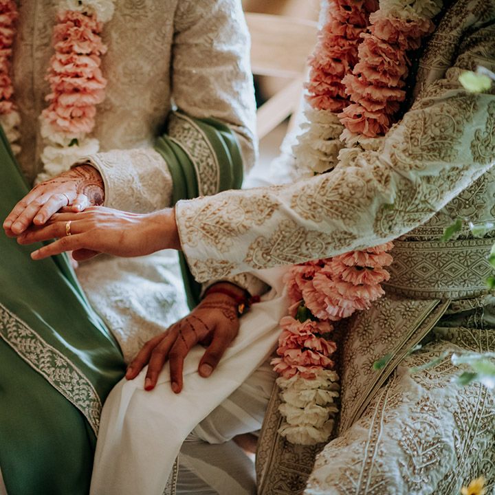 Grooms wearing matching sherwani's with intricate pattern hold their hands above one another during wedding ceremony in barn