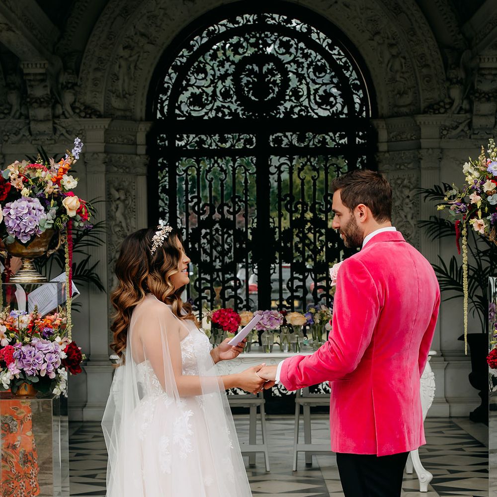 Bride in a tulle wedding dress and sheer bridal cape reads out her wedding vows as she holds onto the groom's hand 