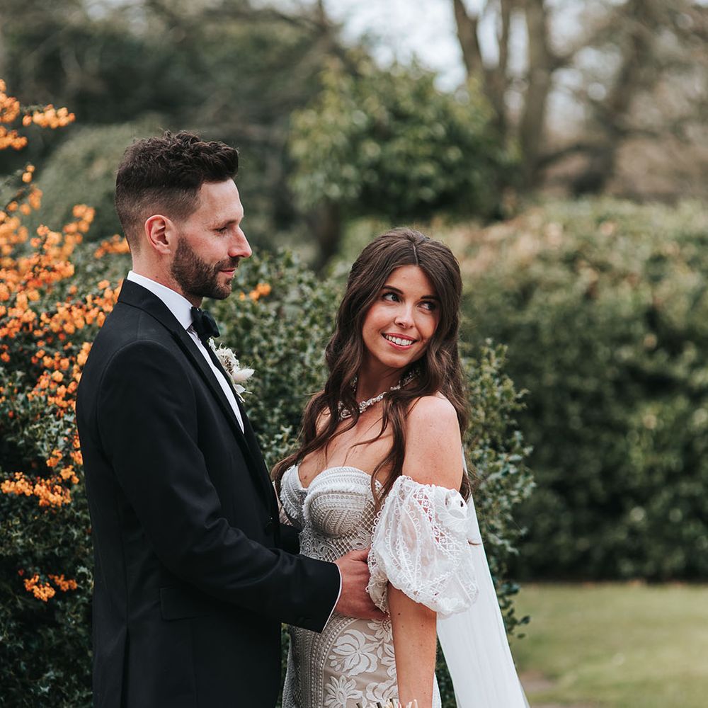 The groom in a black tuxedo gazes at the bride in a fitted boho lace gown with detachable sleeves posing for their couple portraits 