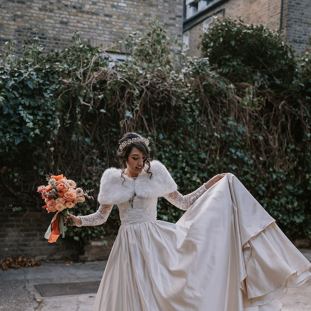 Bride in taffeta material princess wedding dress with long lace sleeves  and cropped faux fur bridal cover up with gold, pearl and emerald bridal crown holding bouquet with light pink poppies, orange garden roses, eucalyptus, Christmas red berry garlands, dried flowers and sage green and orange ribbon