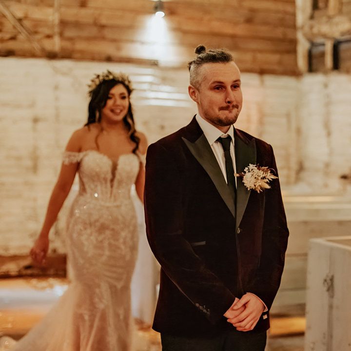 The bride in an off the shoulder lace wedding dress walks up behind the groom in a dark red velvet suit jacket for the first look moment 