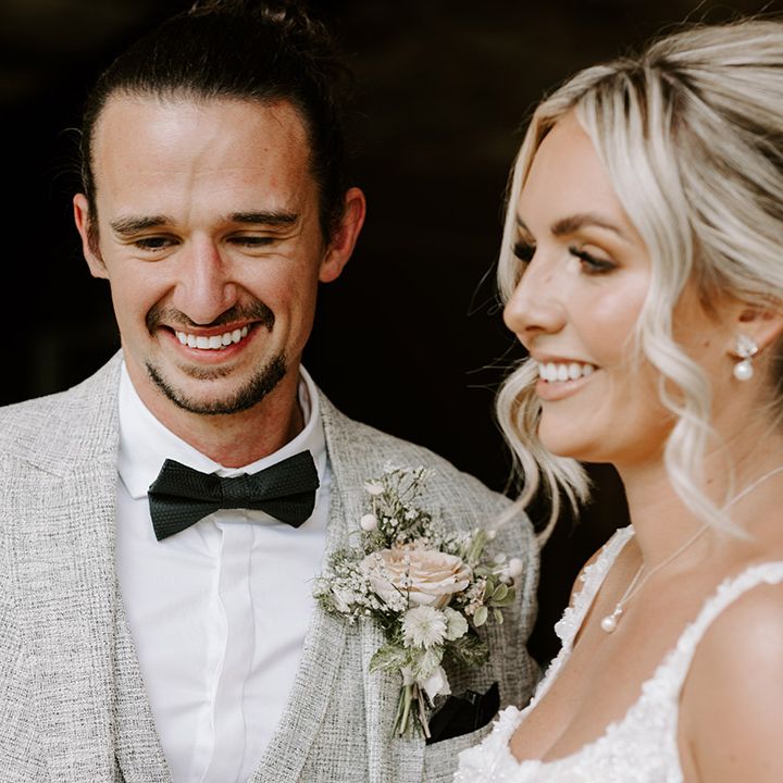Smiling groom in light grey three piece suit and dark bow tie with neutral floral buttonhole with the bride wearing her blonde hair in an updo with a pearl veil, necklace and earrings 
