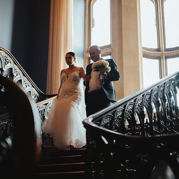 Bride in sleeveless v neck Enzoani wedding dress with tulle finish and church length veil going down the stairs at Grittleton House with father of the bride 