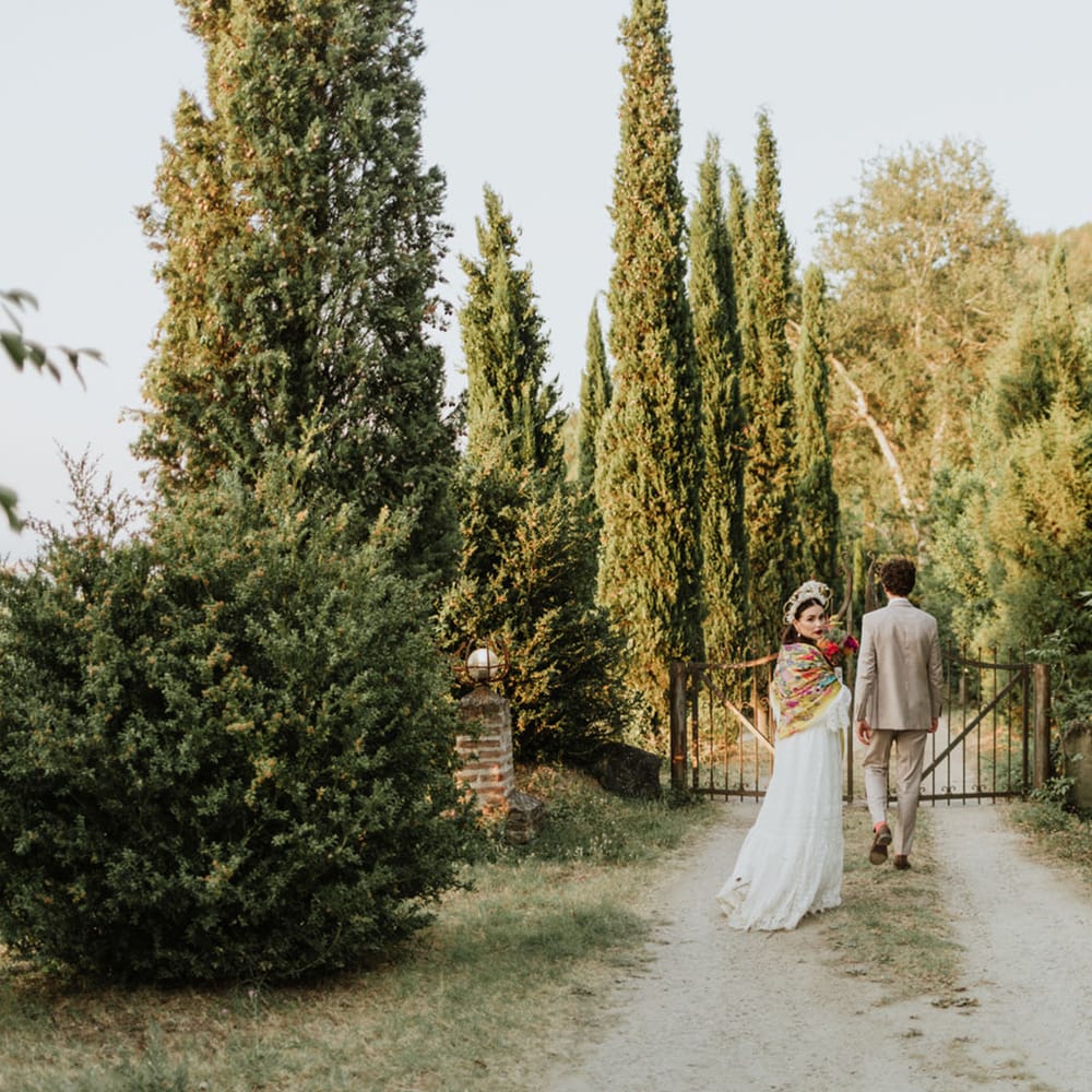 Bride and groom walk through greenery at italian destination wedding wearing white lace wedding dress, white floral structured bridal crown and greige suit