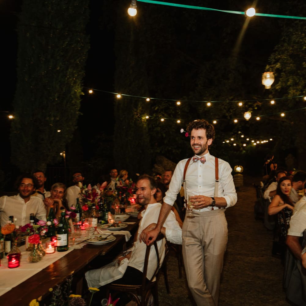 Groom in burnt orange braces holds glass of champagne at wedding reception in Italy