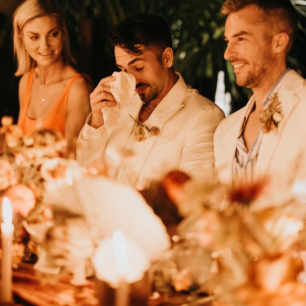 Groom in mens white wedding suit wiping a tear away during wedding speeches at destination wedding 