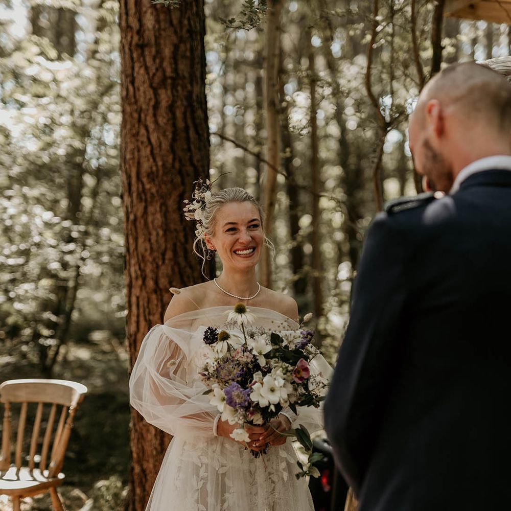 Bride holding purple and white wildflower wedding bouquet with floral lace off the shoulder wedding dress with detachable sleeves smiling up at the groom 