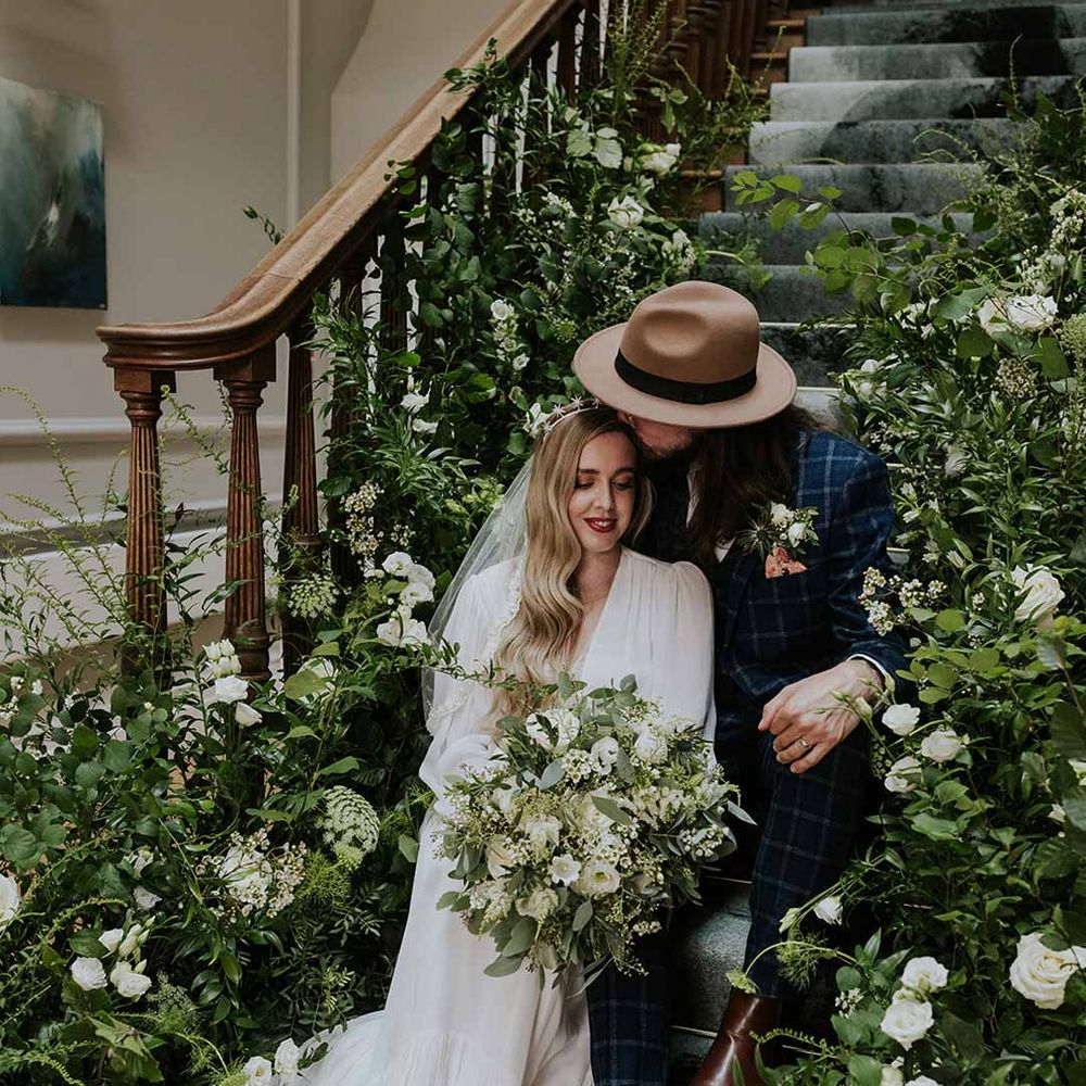 Bride in v-neck long sleeve puff wedding dress with groom in alternative checkered suit and hat sitting on the stairs of Netherbyres House Scottish wedding venue surrounded by white flowers and foliage arrangements 