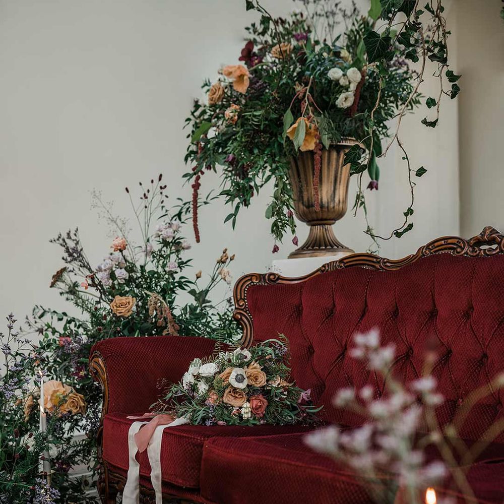 Deep maroon velvet loveseat surrounded by tapered candles and suspended wildflower and foliage arrangements in large gold vases at Buxted Park