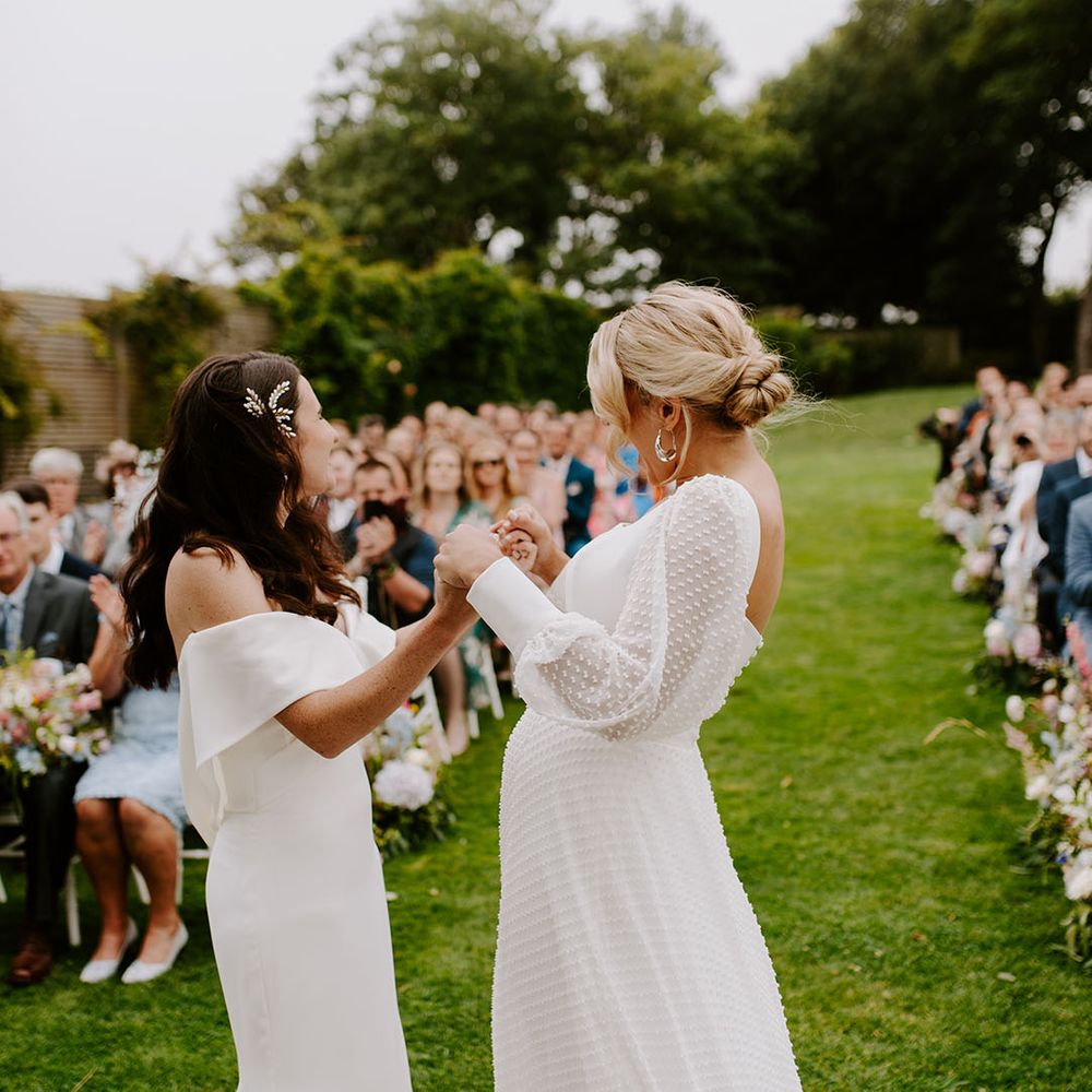 Two brides holding hands together at their same sex outdoor wedding ceremony 