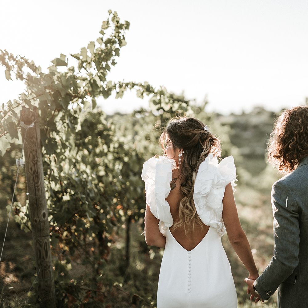 Bride in low back wedding dress with button detail and ruffle sleeves holding her grooms hand in a blue suit in a vineyard in Italy 