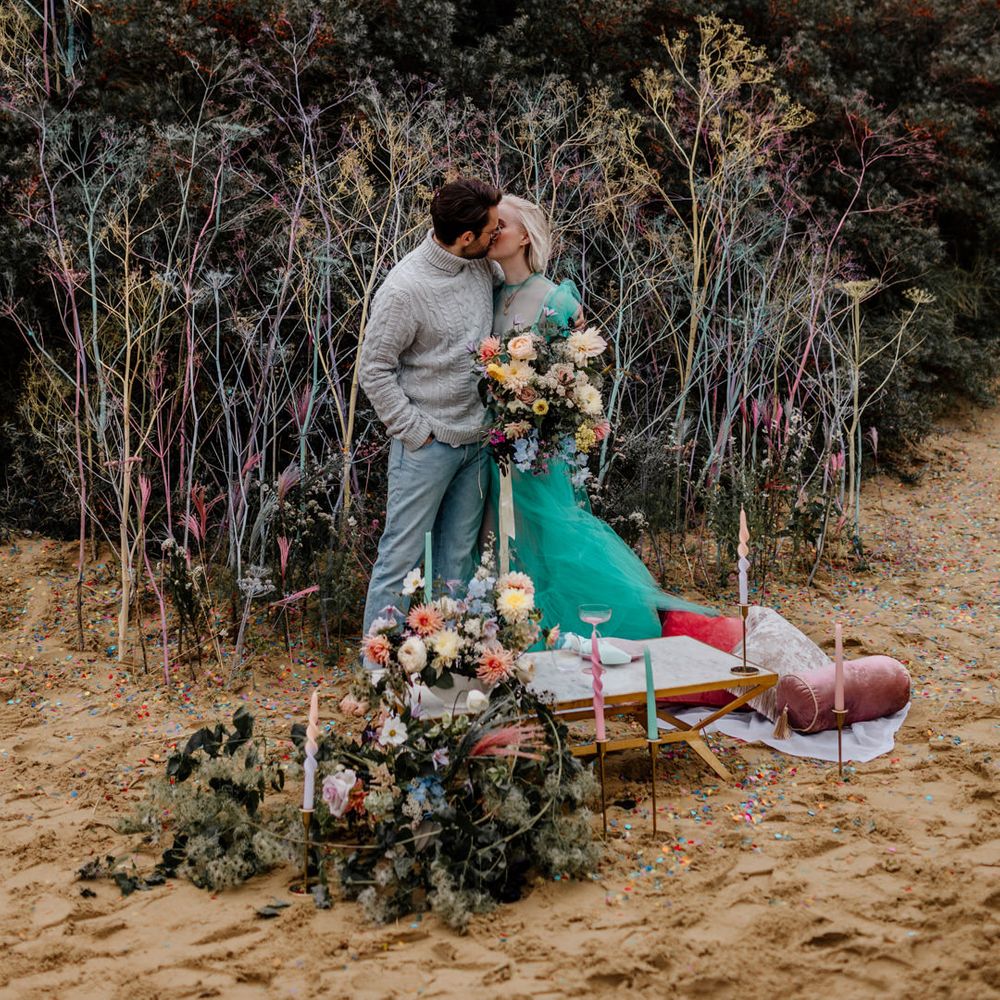 Groom in a grey turtle neck jumper kissing his bride in a green sheer wedding dress at their intimate beach elopement with colourful flowers and picnic area 