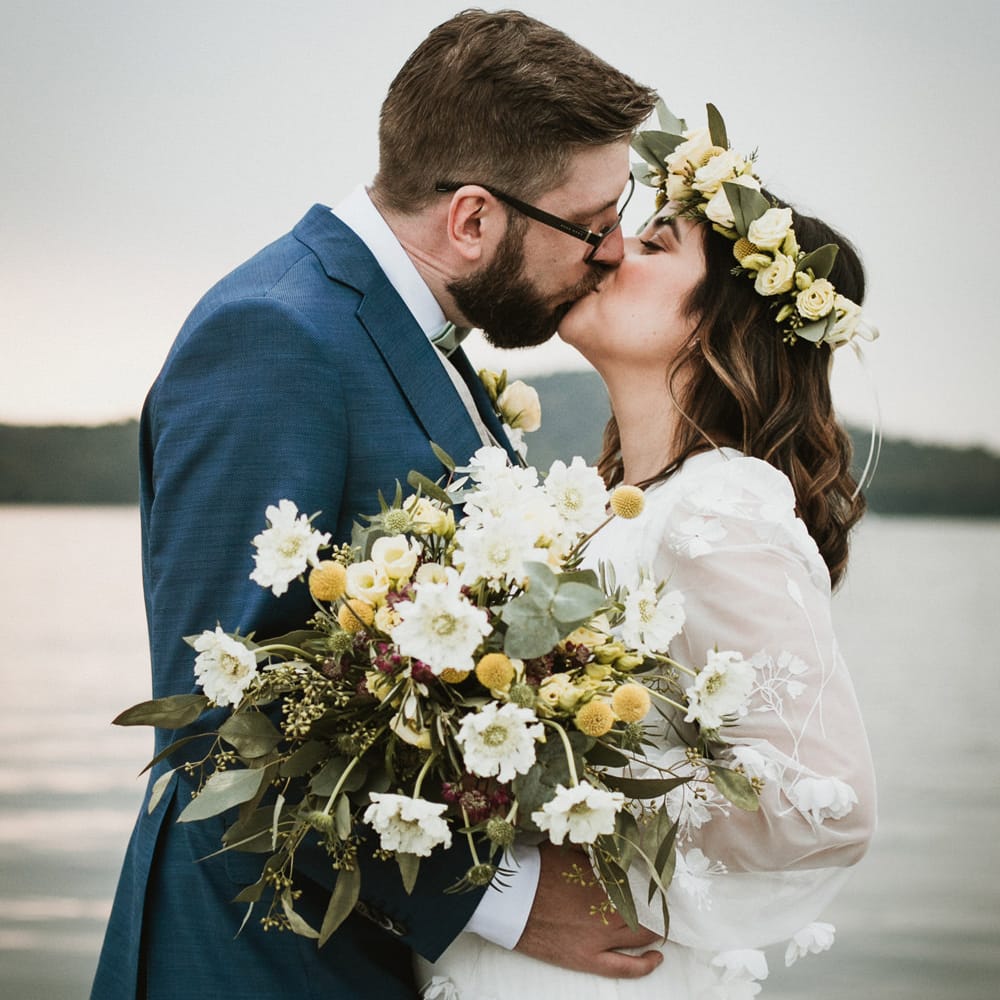 Groom in a navy blue suit kissing his boho bride in a yellow, white and green flower crown holding an unstructured wedding bouquet 