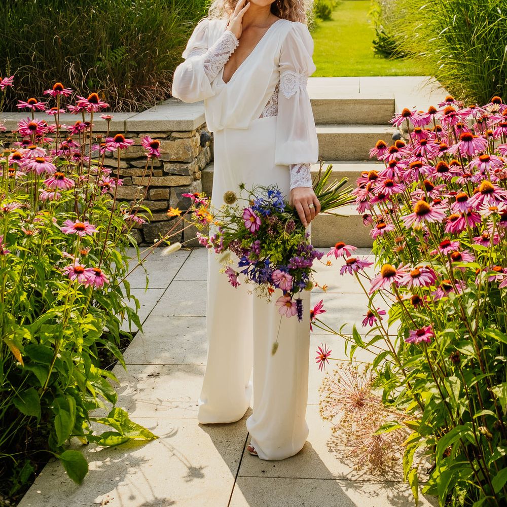 Blonde naturally curly haired bride with bright red lipstick in a jumpsuit with lace cuff sleeve detail holding a wildflower bouquet 