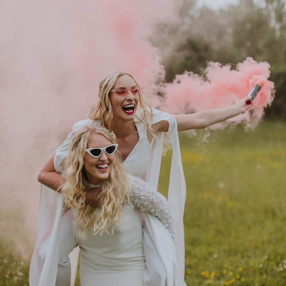 Bride in a short wedding dress giving her bride in a jumpsuit a piggy back as she holds a pink smoke bomb 