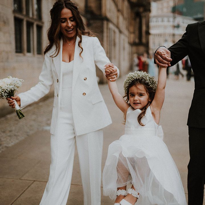 Flower girl in a tulle skirt dress and gypsophila flower crown being swung by her bride and groom parents in Reiss suits 
