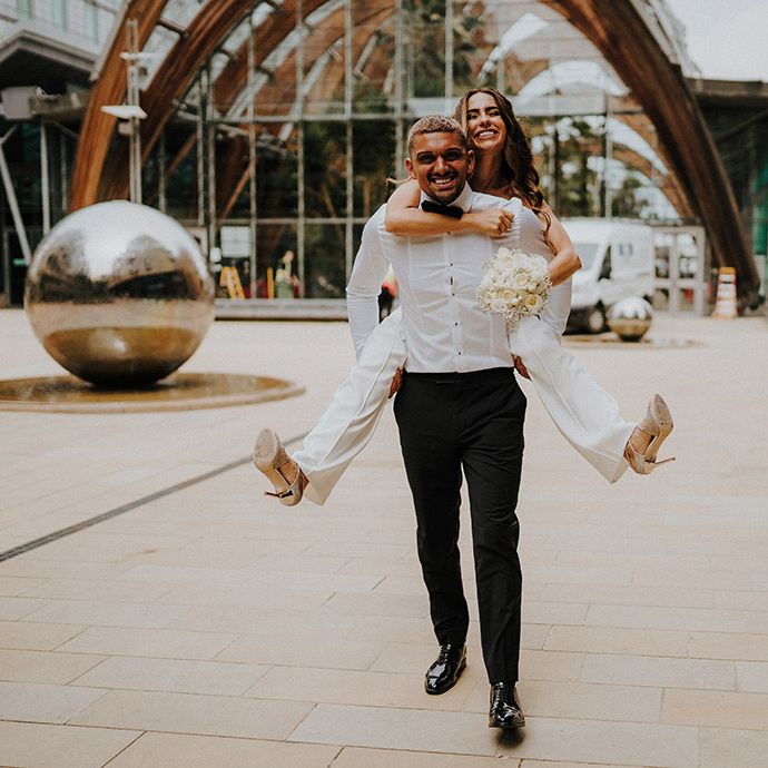 Groom in a white shirt and bow tie earring his bride in a white suit on his back 