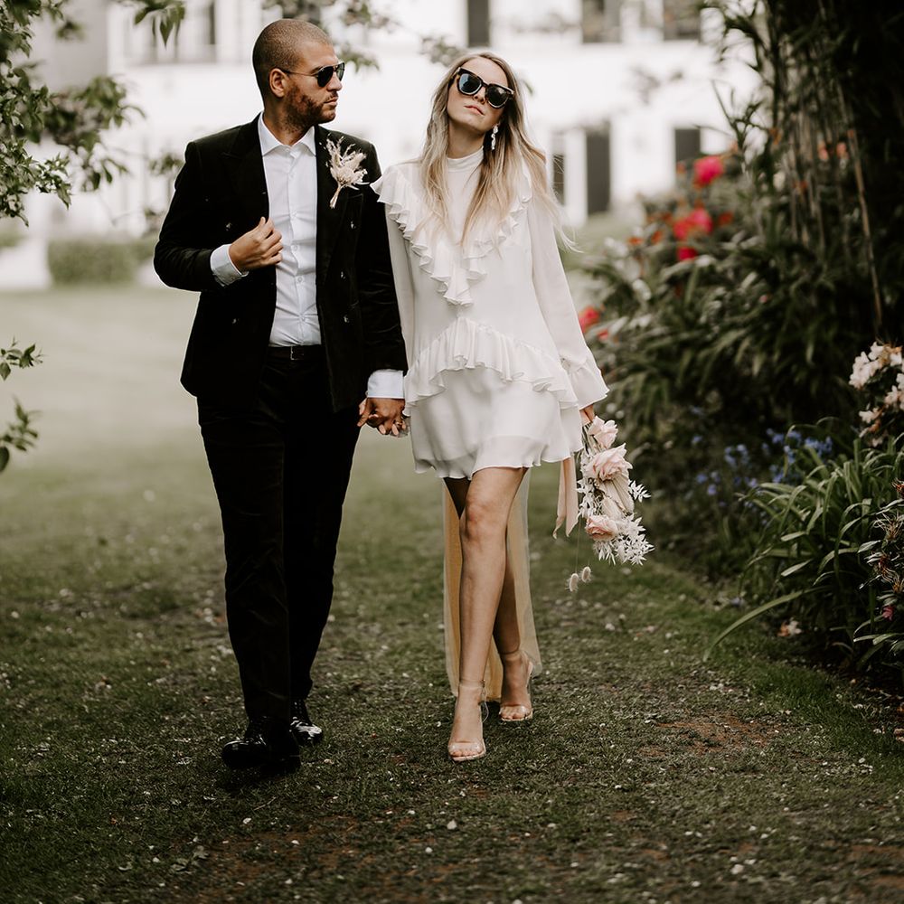 Groom in a black suit with dried flower buttonhole holding hands with his bride in a short wedding dress with ruffle collar in the gardens at Waresley Park Estate 