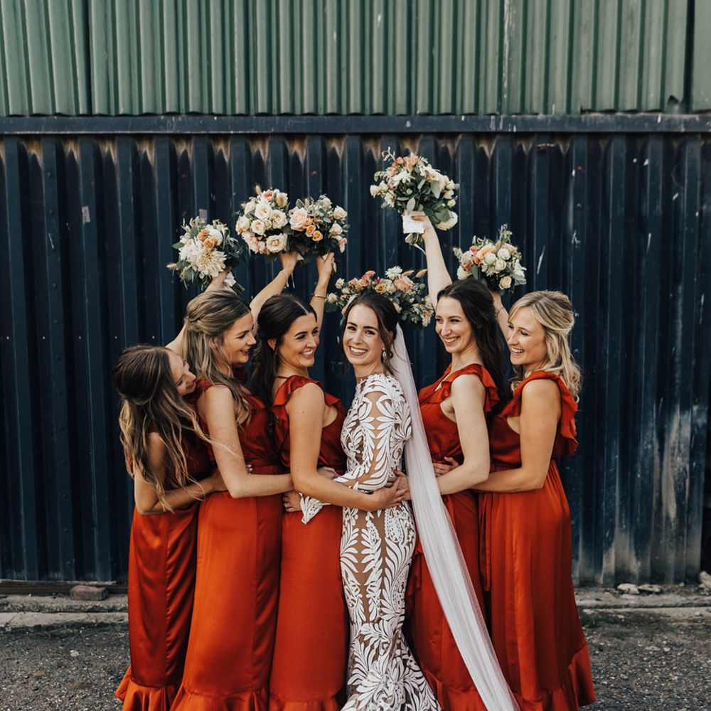 Bridal party with milk churn flower arrangement in the air & silk orange bridesmaid dresses stand with Boho Bride 